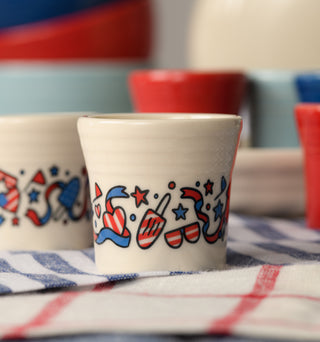 The Fiesta 4th of July 1 1/2 OZ Ceramic Shot Glass, featuring red, white, and blue patriotic stars and hats, rests on a blue and white checkered cloth with similarly themed tableware blurred in the background.