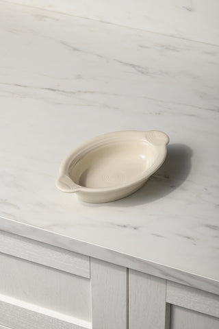 A Fiesta 9 1/8 Inch Oval Individual Casserole, 13 oz, in cream ceramic with handles sits on a white marble countertop with gray veining.