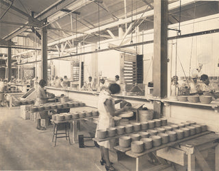 Workers in a factory, mostly women, stand at long tables handling rows of large round wheels, possibly cheese, in an industrial setting with high ceilings and exposed beams.