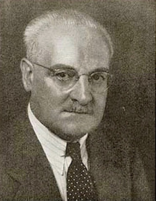 Black and white portrait of an older man with glasses, short white hair, and a mustache, wearing a suit, white shirt, and polka-dot tie, looking directly at the camera.