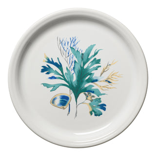 The Fiesta Retired Coastal Shores Bistro Coupe 9 Inch Luncheon Plate is a white ceramic plate with a blue seashell and green, blue, and yellow botanical accents. This lead-safe china is dishwasher safe for easy cleaning.