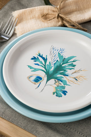 The Fiesta Retired Coastal Shores Bistro Coupe 9 Inch Luncheon Plate, featuring blue and green botanical and seashell motifs, rests on a light blue charger over a gray placemat beside a twine-tied beige napkin and silverware. Made in the USA with lead-safe china.