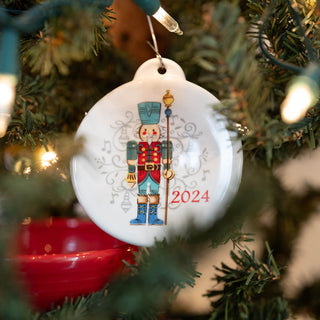 A Fiesta Retired Nutcracker Guardian 2024 Ornament hangs on a festive, decorated tree with twinkling lights. This white ornament features a colorful nutcracker and is proudly made in the USA.
