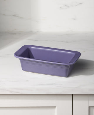 A Fiesta Lavender 10 3/4 Inch x 5 3/4 Inch Loaf Pan Baker (1.4 Quarts), made in the USA and oven safe, rests on a white marble countertop with light gray cabinets and a white marble backsplash in the background.