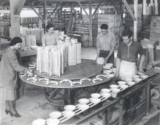 Four people in a factory arrange ceramic plates on a rotating table and conveyor belt, surrounded by stacks of dishes and shelves filled with pottery. The factory has a rustic, open wooden structure.
