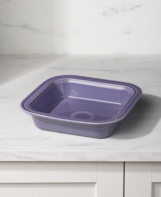 The Fiesta Lavender Square 9x9 Inch Baker, holding 2 quarts, rests on a white marble countertop with subtle gray veining against a white background. Proudly made in the USA.