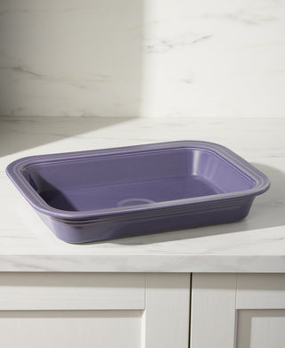 A Fiesta Lavender Rectangular 9x13 Inch Baker (3 Quart) sits on a white marble countertop with light gray cabinets below and a light marble backsplash. This oven safe bakeware is proudly made in the USA.