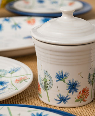 The Fiesta Retired Breezy Floral 13 OZ Mini Canister (5 5/8 Inch) is a white, lead-safe china canister with a lid and colorful floral designs, shown on a woven mat with matching plates. Dishwasher safe for easy cleaning.