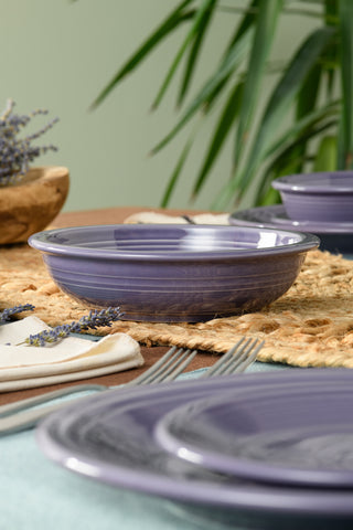 A close-up of a table setting featuring Fiesta's Lavender Classic Rim 8 3/8 Inch Pasta Bowl (34 oz) on a woven placemat with silver forks, a beige napkin, and sprigs of lavender amid green plants in the background.