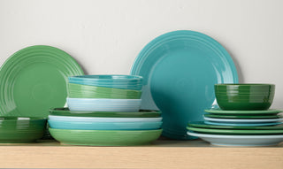 A stack of green, blue, and turquoise ceramic plates and bowls arranged on a light wooden shelf against a white wall. The dishes are organized in neat, overlapping piles.