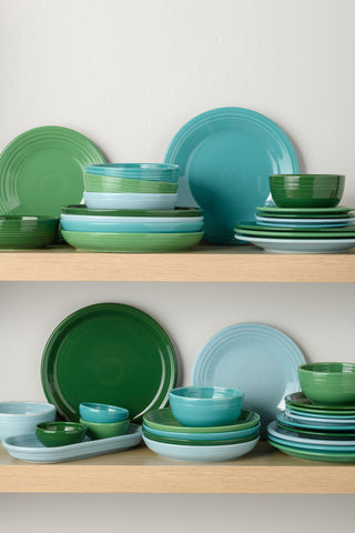 Two wooden shelves display neatly stacked ceramic dishes in shades of green, blue, and teal, including plates, bowls, and saucers, arranged against a plain white wall.