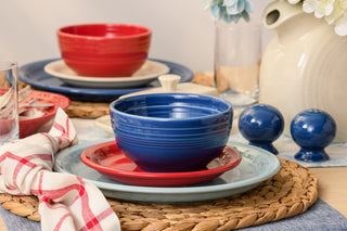 A modern table is set with Fiesta's Mixed Americana Bistro Coupe 12-Piece Dinnerware Set—blue bowls, red and light blue plates, plus a red bowl on a beige plate. Woven placemats, napkins, shakers, and a white floral pitcher complete the look. Dishwasher safe.
