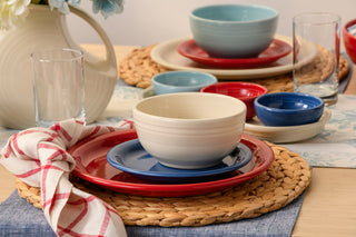 A neatly set table features the Fiesta Mixed Americana Bistro Coupe 12-Piece Dinnerware Set, Service for 4—modern pieces in red, blue, and cream—arranged with woven placemats, clear glasses, a floral pitcher, and a red-and-white checkered napkin.