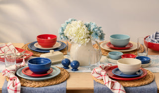 A dining table set for four with red, blue, and cream dishes, woven placemats, clear glasses, and checkered napkins. A white pitcher with white and blue flowers serves as the centerpiece.