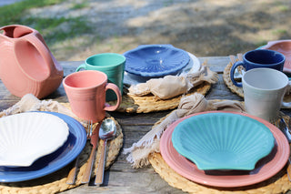 A rustic outdoor table is set with pastel plates, Fiesta Retired Shell Shaped Plate 8 1/4 OZ, mugs, a large pink pitcher, woven placemats, beige napkins, and lead-safe china for a colorful beach-themed look.
