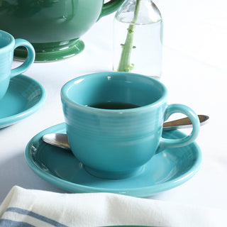 A Fiesta Retired Classic 7 3/4 OZ Tea Cup with matching saucer, made from lead-safe china, holds a spoon and dark liquid on a white tablecloth next to a clear bottle containing a green stem.