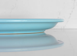 A close-up side view of a light blue ceramic plate on a reflective white surface, with a white marble-patterned background.