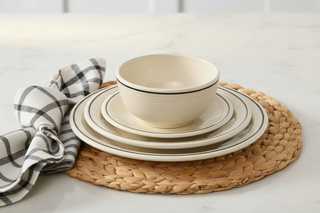 A stack of cream-colored dishes and a bowl featuring black rims from the Fiesta Factory Direct Black Band Classic Bistro 3-Piece Dinnerware Set, Service For 1, sits on a woven placemat beside a folded black-and-white checkered napkin on a marble surface.