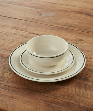 A three-piece ceramic dinnerware set consisting of a bowl, a saucer, and a plate, stacked neatly on a rustic wooden table. The dinnerware is off-white with thin black double-lined borders.