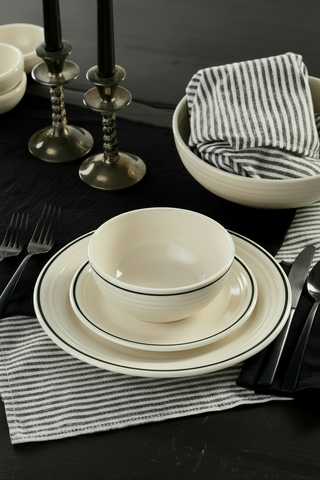A place setting with white dishware, including a Fiesta Black Band Classic Rim 9 Inch Luncheon Plate, black utensils, and striped black-and-white napkins on a dark table, featuring candles in metal holders.