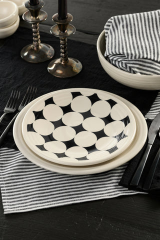 A table setting featuring the Fiesta Black Kaleido Circles Classic Rim 9 Inch Luncheon Plate, a striped napkin, sleek silver cutlery, and elegant candle holders arranged on a black tabletop highlights the modern linen dinnerware style. The distinctive geometric design of the Fiesta plate adds a striking visual element to this chic presentation.