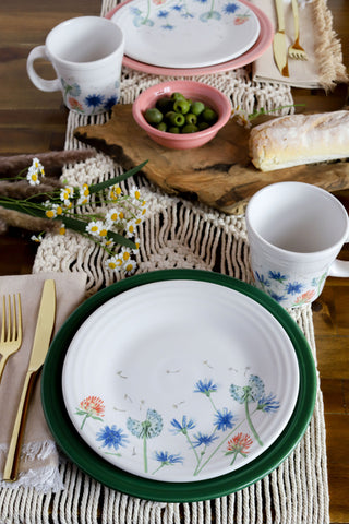 A wooden table set with Fiesta Retired Breezy Floral Classic Rim 9 Inch Luncheon Plates, gold cutlery, mugs, a bread loaf, green olives, and a white crochet table runner adorned with small white flowers.