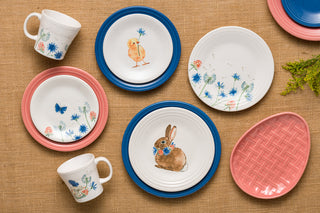 The Fiesta Retired Breezy Floral Easter Classic Rim 9 Inch Luncheon Plate features blue flowers, butterflies, a chick, and a bunny with pink accents on a burlap surface. Dishwasher safe for easy cleaning. Includes a pink oval dish with wicker-like texture.