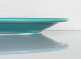 A side view of a turquoise plate on a reflective surface, showing its thin edge and subtle reflection against a white background.