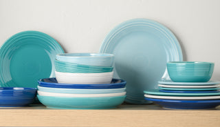 A stack of assorted ceramic dishes, bowls, and plates in shades of blue, teal, and white, arranged neatly on a light wooden shelf against a plain white wall.