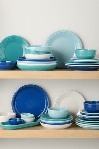 Two wooden shelves display neatly stacked ceramic plates and bowls in shades of blue, teal, and white, arranged in an organized and visually pleasing manner against a plain white wall.