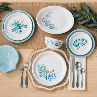 A wooden table is set with the Fiesta Retired Bistro Coupe 3-Piece Place Setting, Service for 1 Coastal Shores, featuring blue and green floral dinnerware, silver cutlery, shell-shaped plate, woven placemat, green leaves, and a burlap runner.