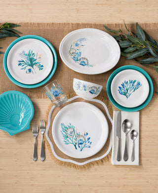 A top-down view of a table set with Fiesta’s Retired Coastal Shores 11 5/8 Inch Medium Oval Serving Platter, white vitrified china with blue and green botanical designs, turquoise plates, silver cutlery, glassware, and napkins on a bamboo surface.