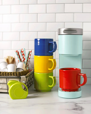 A pastel-blue single-serve coffee maker, made in the USA, sits on a marble counter by retired Fiesta 16 OZ Stackable Mugs in blue, yellow, green, and red. Lead-safe china and coffee accessories are nearby. White subway tile backsplash.
