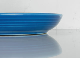 A close-up side view of a blue ceramic plate, shown against a white background with its reflection visible on a shiny surface.
