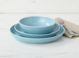 A set of three light blue ceramic dishes—one bowl, one salad plate, and one dinner plate—stacked on a white marble surface, with a beige napkin, fork, and knife beside them.
