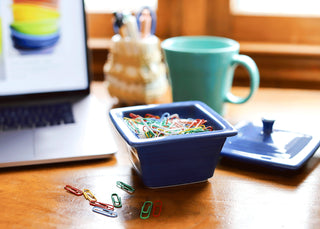 A Retired Fiesta Covered Box (4 5/8" x 3 3/4") by Fiesta sits on a wooden desk with colorful paperclips, beside a laptop, turquoise mug, and scattered clips. The background has blurred office supplies and a window.