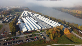 A large industrial building with a white roof sits beside a river, surrounded by trees with autumn foliage. Parking lots with many cars are in front, and fog covers the river in the background.
