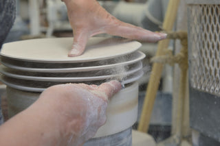 A person shapes and smooths the edges of stacked ceramic plates on a pottery wheel, with clay dust on their hands and fingers.