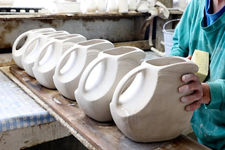A person shapes or cleans a row of large, unfinished ceramic vases on a workbench in a pottery studio, using a sponge on one vase. The vases are uniform and in various stages of drying.