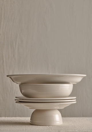A stack of white ceramic dishes, including plates and bowls, is arranged on a light-colored textured surface against a neutral, textured background.
