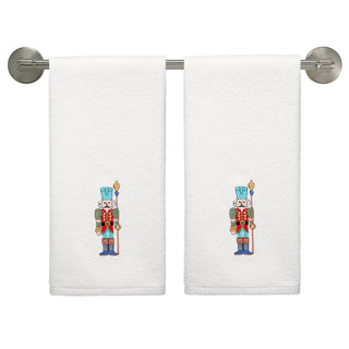 The Fiesta Nutcracker The Guardian Embroidered Hand Towel Set of 2 features white towels with nutcracker soldiers in red, blue, and black uniforms holding staffs, perfect for holiday decor.