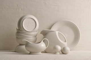 A set of white ceramic dishes, including stacked bowls, a pitcher, a gravy boat, a plate, and salt and pepper shakers, arranged against a textured off-white background.