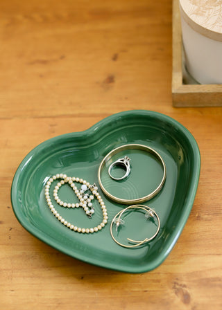 A retired Fiesta 7 1/2 inch medium heart-shaped bowl (17 oz) in green holds bracelets, a beaded necklace, earrings, and a ring on a wooden surface next to a small box.