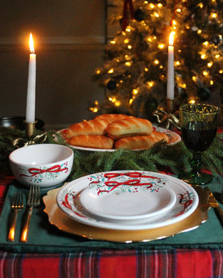 A festive holiday table is set with the Fiesta Holly Berry Christmas Garland Classic Bistro Coupe 3-Piece Place Setting, a bowl, green wine goblet, silverware, bread rolls, candles, and evergreen branches before a lit Christmas tree.
