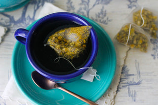 A Fiesta Retired Classic 7 3/4 OZ Tea Cup with chamomile tea sits on a matching saucer, bronze spoon beside it. Two tea bags rest on a white lace-edged napkin atop a patterned tablecloth. Made from lead-safe china.