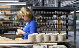 A woman in a blue shirt shapes a clay pot by hand at a pottery workshop, with finished and unfinished clay pots arranged on shelves and tables around her.