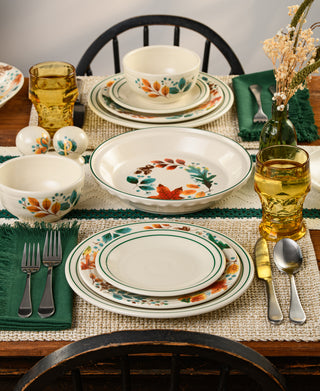 A set dinner table with floral-patterned plates, bowls, and glasses, green napkins, silver cutlery, and a vase with dried flowers on a woven placemat over a wooden table.