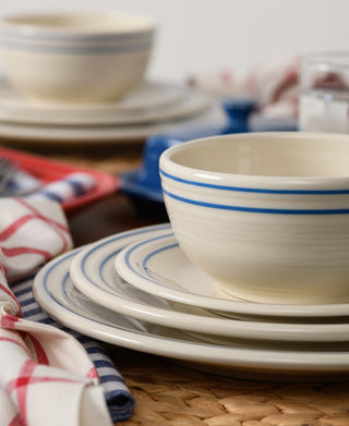 A close-up of stacked Fiesta Americana Lapis Band Classic Bistro 3-Piece Place Setting, Service For 1, featuring cream dishes with blue stripes on a woven placemat. Red, white, and blue checkered napkins add charm to this USA-made set.