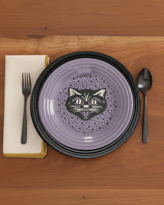 The Black Cat Lavender Classic Rim 9 Inch Luncheon Plate from Fiesta, crafted from vitrified china and made in the USA, features a purple hue with a black cat face and stars design. It sits atop a black charger on a wooden table, with a fork, knife, and napkin arranged to the left and a spoon to the right. This dishwasher-safe plate adds charm to any table setting.