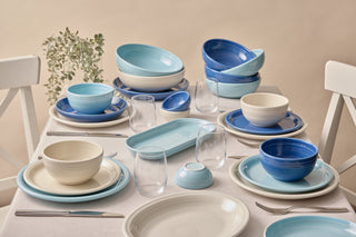 A dining table set with blue and white ceramic plates, bowls, and glassware, arranged neatly with forks and knives. A small potted plant sits in the corner, creating a fresh and inviting atmosphere.